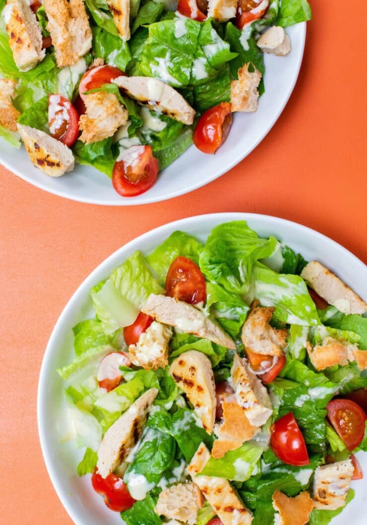 Vegetarian Caesar salad with Quorn chicken pieces, lettuce, tomatoes, croutons and Caesar dressing in 2 white bowls on an orange background.