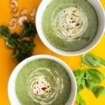 2 bowls of broccoli and kale soup with a swirl of cashew cream with kale, cashew and basil shattered over the yellow background.
