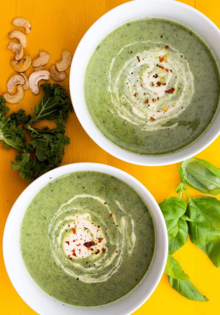 2 bowls of broccoli and kale soup with a swirl of cashew cream with kale, cashew and basil shattered over the yellow background.