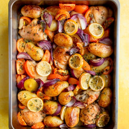 Lemon & Thyme Chicken thigh Tray Bake with new potatoes, red onions, tomatoes and topped with lemon round slices in a baking tray.