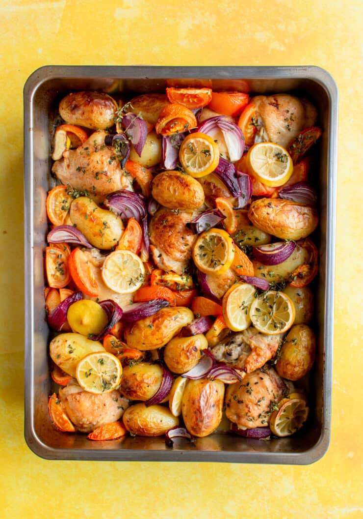 Lemon & Thyme Chicken thigh Tray Bake with new potatoes, red onions, tomatoes and topped with lemon round slices in a baking tray.