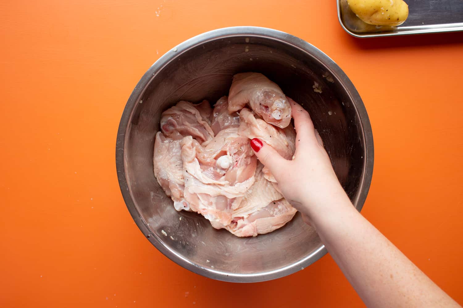 Chicken thighs in a metal bowl being mixed by hand on an orange background.