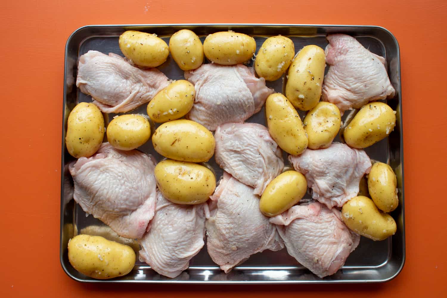 Chicken thighs and new potatoes on a stainless steel baking tray on an orange background.