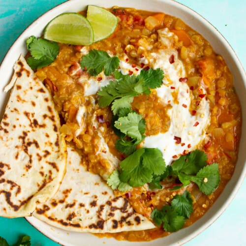 A bowl of Red Lentil Dahl with Coconut Milk with 2 slices of naan bread and topped with fresh coriander and lime wedges.