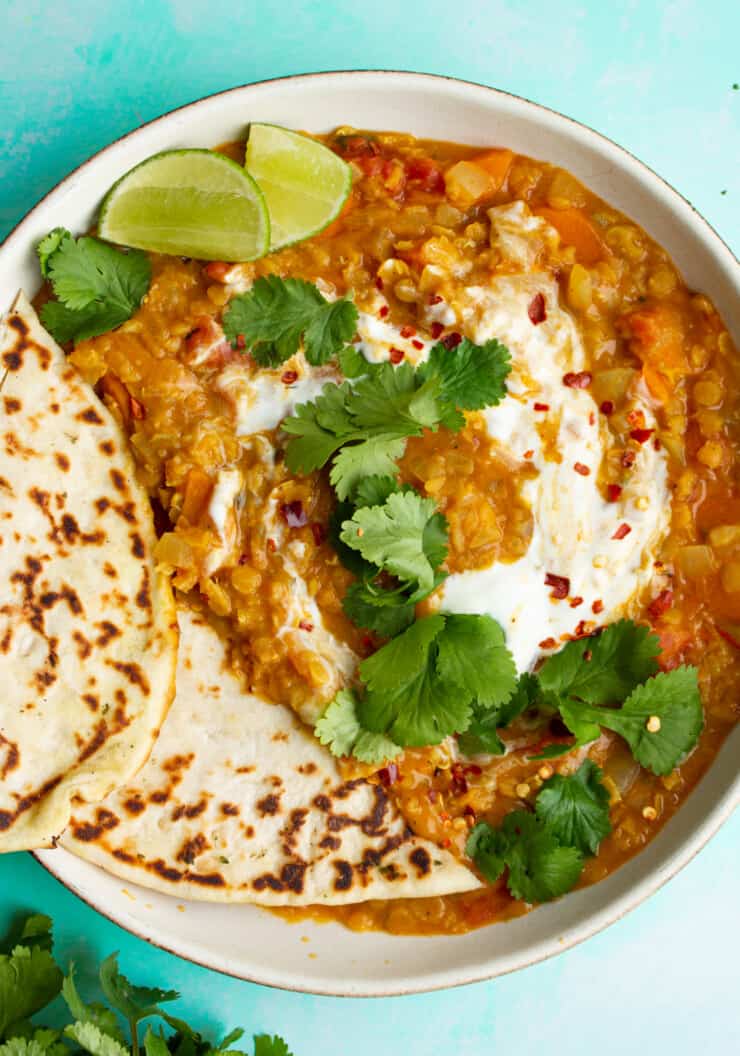 A bowl of Red Lentil Dahl with Coconut Milk with 2 slices of naan bread and topped with fresh coriander and lime wedges.