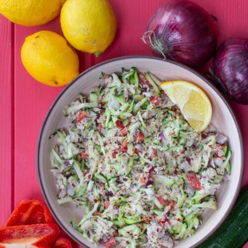 Overhead shot of bowl of tuna salad with grated courgette, and lemons and red onions on display near bowl