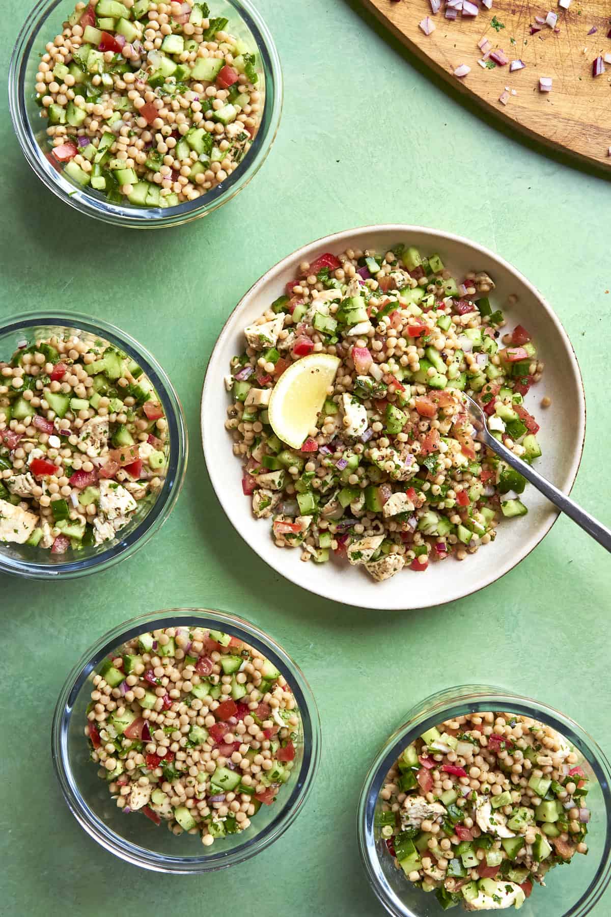 Five glass bowls and one plate contain a colorful couscous salad with diced cucumber, tomato, parsley, and feta cheese. A lemon wedge and spoon sit on the plate. A cutting board with chopped onions is nearby on a green surface.