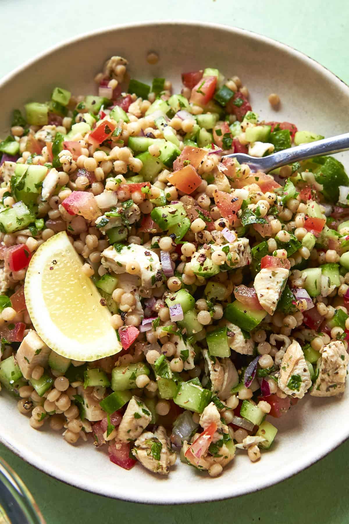 A bowl of couscous salad with diced cucumbers, tomatoes, onions, chicken pieces, fresh herbs, and a lemon wedge, served with a fork.