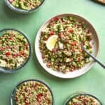 A plate and four bowls of couscous salad with diced cucumber, tomato, herbs, and feta cheese, garnished with a lemon wedge, are arranged on a green table. A fork rests on the main plate.
