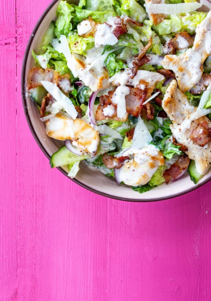 Caesar salad in a bowl with lettuce, chicken and bacon and dressing on a pink background.