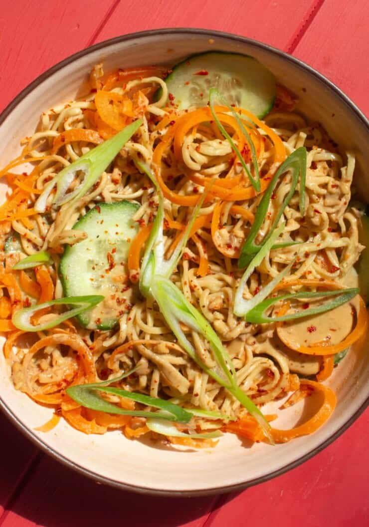 Peanut noodle salad with sprialised carrots, sliced cucumber and spring onion in a bowl on a pink background.