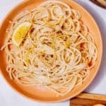 Creamy spaghetti on a plate with lemon rind and a wedge of lemon and a wooden chopping board with a slice of lemon on a board on a white background.