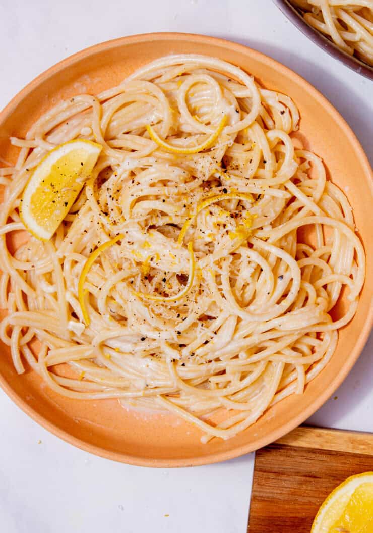 Creamy spaghetti on a plate with lemon rind and a wedge of lemon and a wooden chopping board with a slice of lemon on a board on a white background.