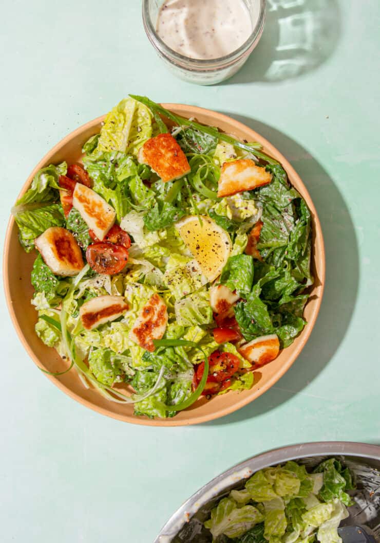 A bowl of salad with golden browned halloumi, lettuce, lemon wedges and tomatoes next to a large bowl with salad and the dressing.