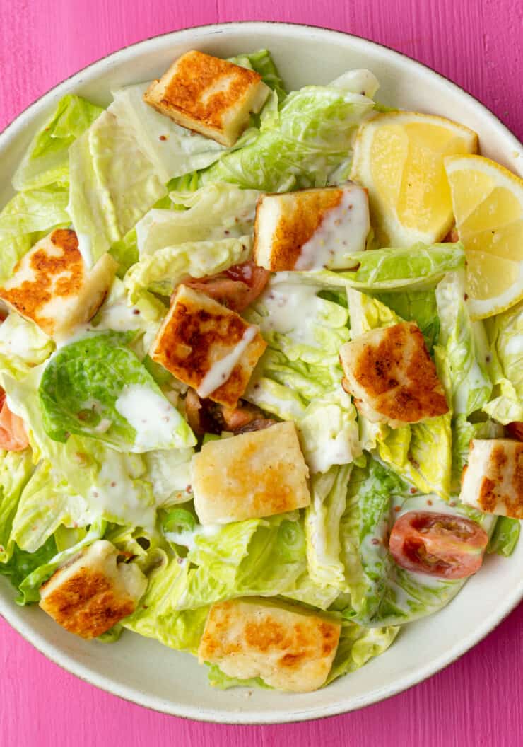 Vegetarian Caesar salad in a bowl with lettuce, golden brown croutons, cherry tomatoes, spring onions and dressing on a pink background.