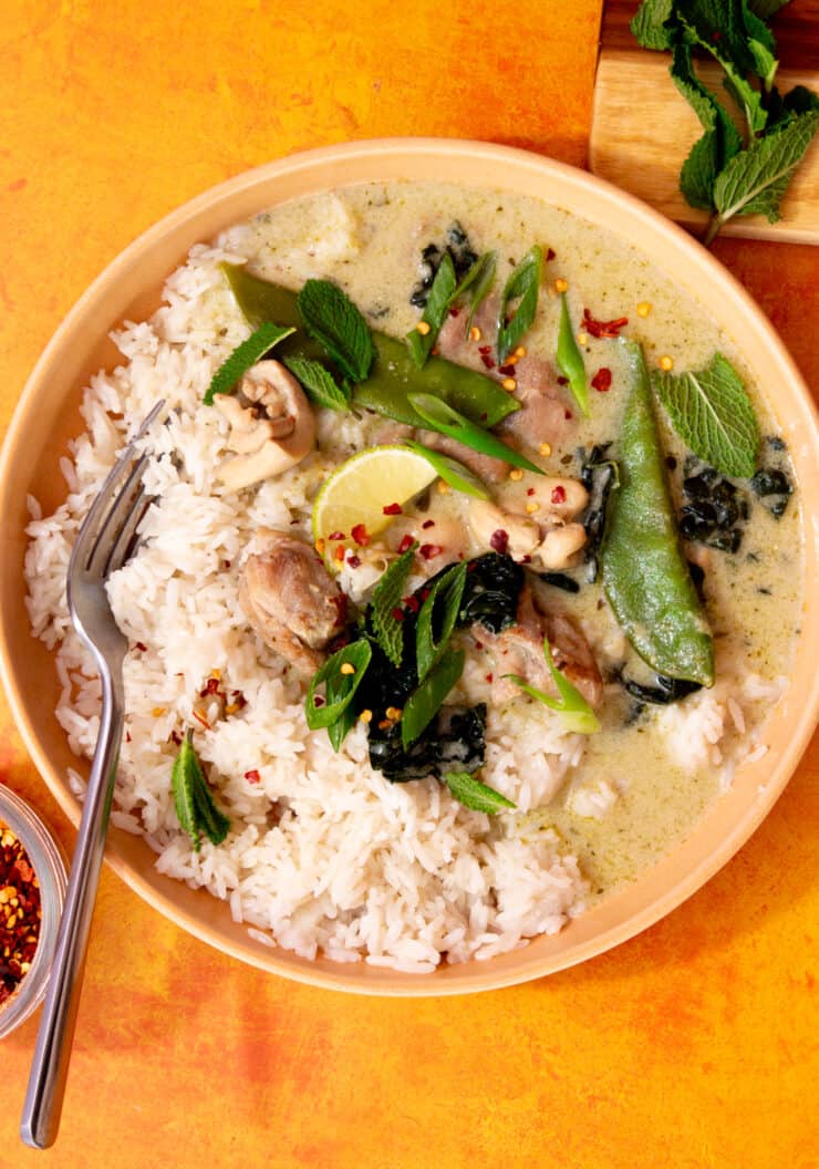 Green curry with mange tout, greens and lime wedges in a bowl with rice in. bowl.