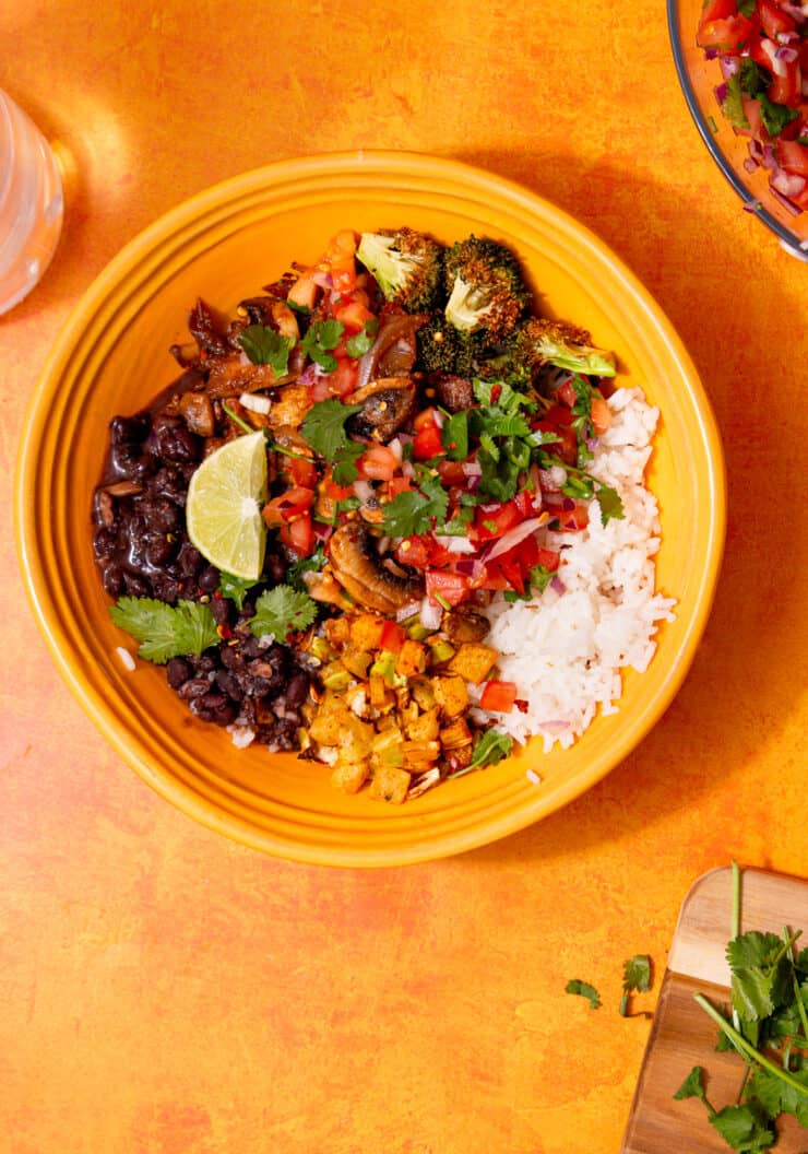 A vegetable burrito bowl with blackens, mushrooms, rice, chopped tomatoes & onion with a lime wedge on an ornage background.
