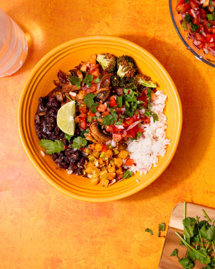 A vegetable burrito bowl with blackens, mushrooms, rice, chopped tomatoes & onion with a lime wedge on an ornage background.