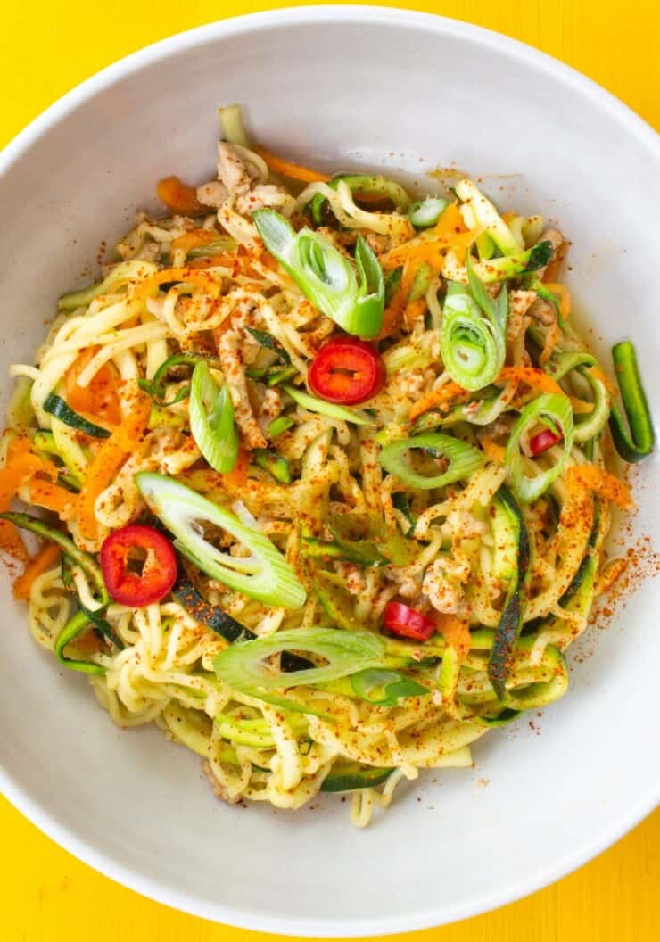 A white bowl of stir fry noodles with pork topped with sliced spring onion and red chillis on a yellow background.