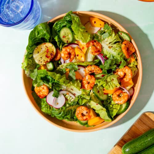 Golden browned king prawns in a bowl of salad with sliced cucumber, radished and red onion next to a blue glass with water.
