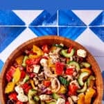 A wooden bowl filled with Greek Chicken salad, featuring colorful vegetables and feta cheese, sits on a blue and white tiled surface. Text above reads Perfect for Meal Prep - Greek Chicken Salad.