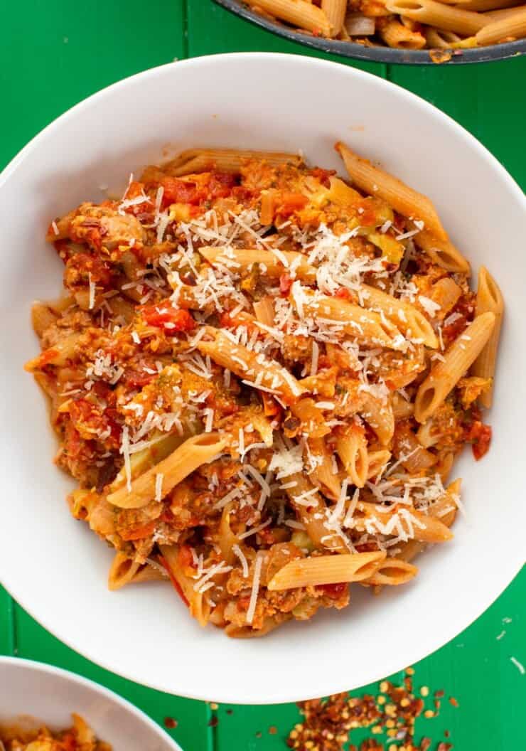 A white bowl filled with penne pasta with tomato, sausage and grated parmesan on a green background.