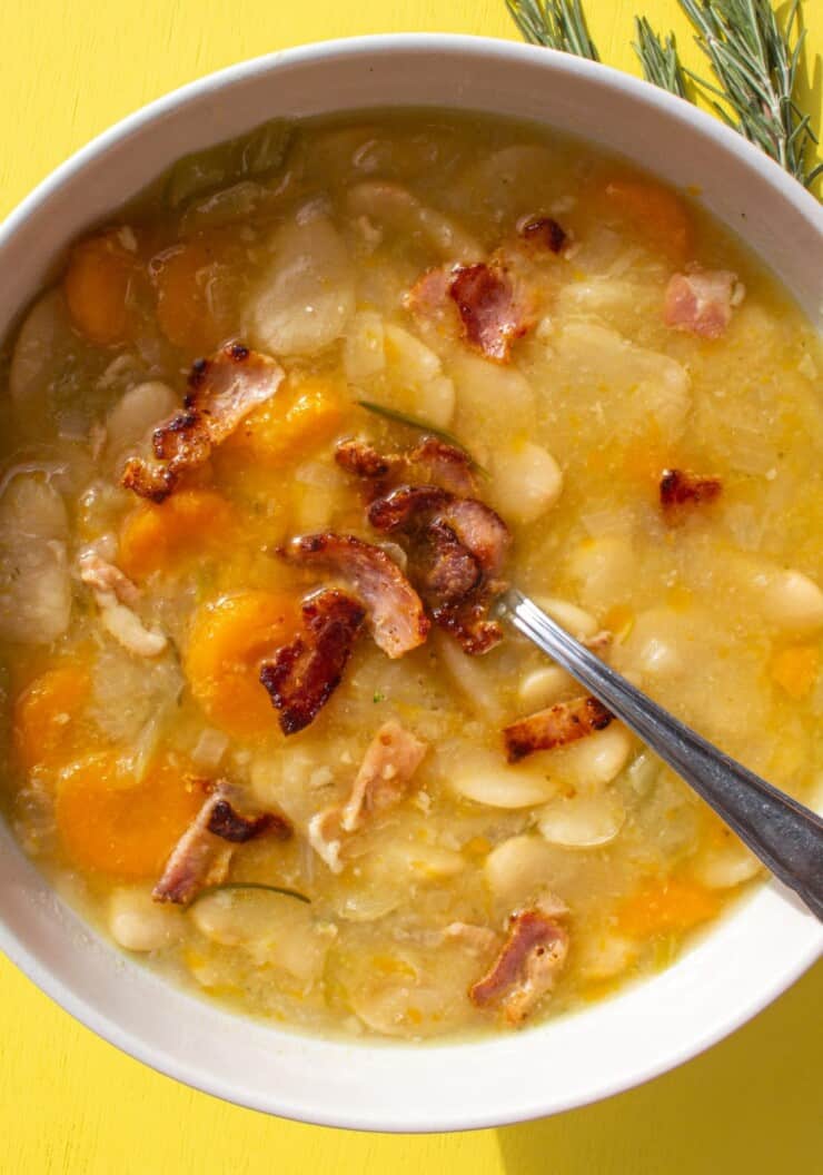A white bowl filled with slow cooker white bean soup with carrots and celery with a crispy bacon topping on a yellow background.