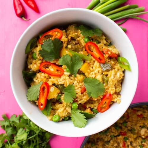 Vegan Egg Fried Rice in a white bowl topped with red chillies and coriander with loose spring onions and coriander on a pink background.