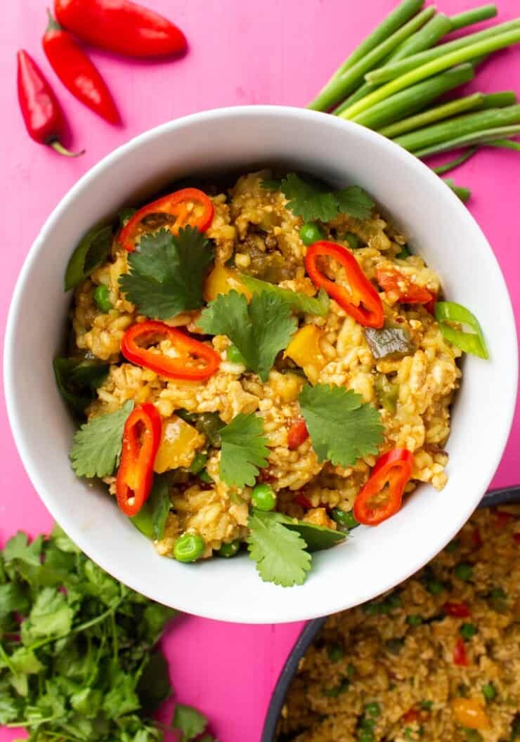 Vegan Egg Fried Rice in a white bowl topped with red chillies and coriander with loose spring onions and coriander on a pink background.