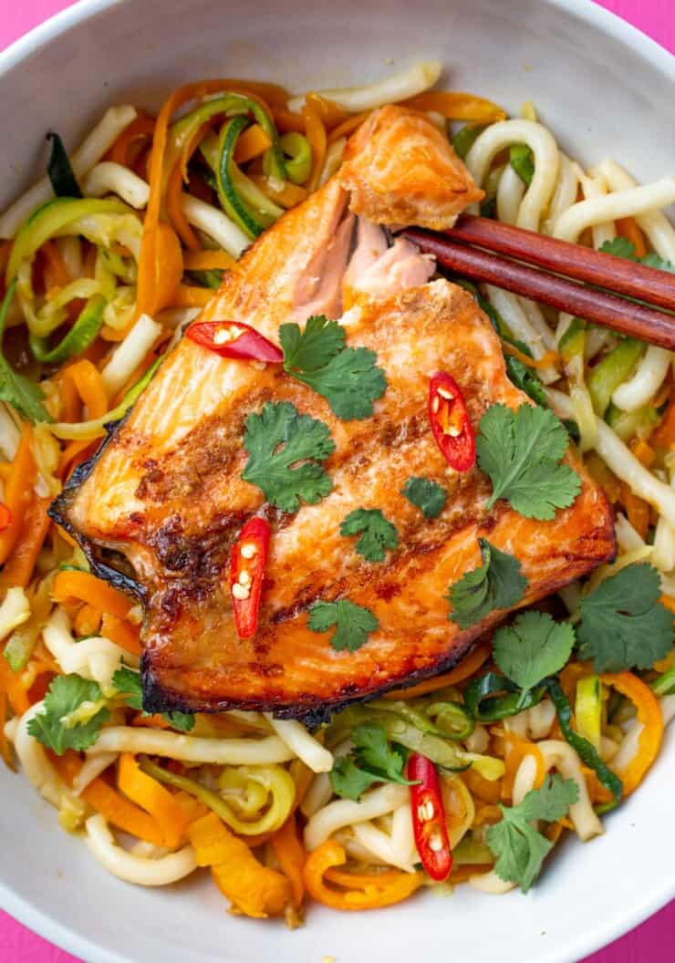 Salmon with chop sticks on a bed of noodles with spiralised courgettes and carrots topped with coriander and red chilli slices.