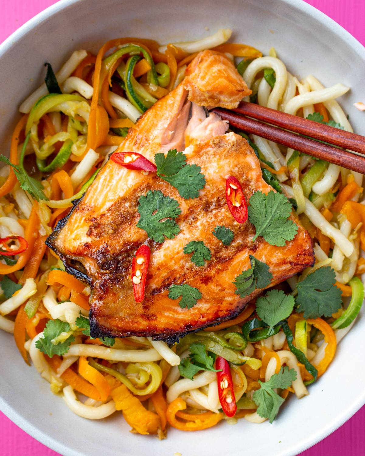 Salmon with chop sticks on a bed of noodles with spiralised courgettes and carrots topped with coriander and red chilli slices.