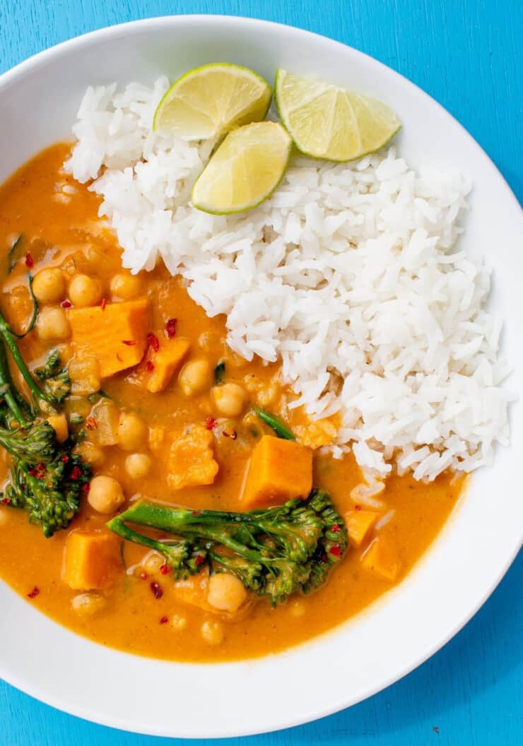 Chickpea and Sweet Potato Curry with broccoli stems served with white rice and topped with lime wedges in a white bowl on a blue background.