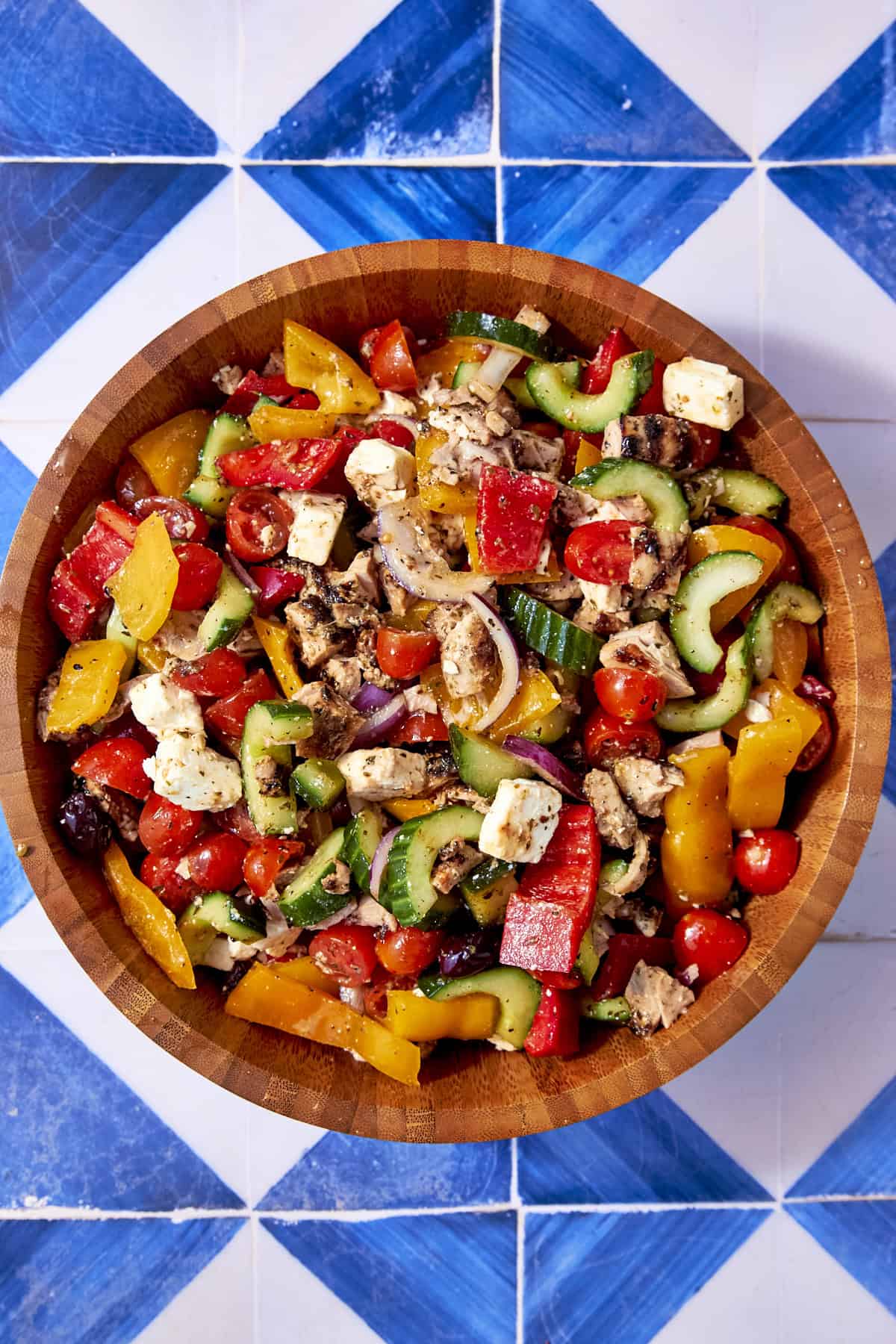 A wooden bowl filled with colorful Greek chicken salad, including tomatoes, cucumbers, red onions, bell peppers, olives, and feta cheese cubes, sits on a blue and white tiled surface.