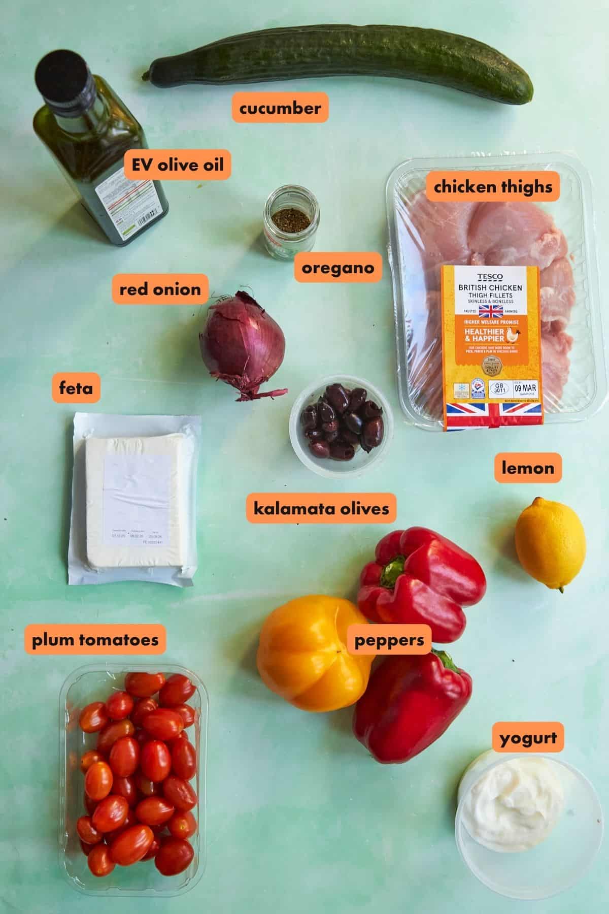 Ingredients for a Greek Chicken salad: chicken thighs, cucumber, red onion, feta cheese, kalamata olives, lemon, plum tomatoes, bell peppers, Greek yogurt, oregano, and extra virgin olive oil arranged on a light green surface.