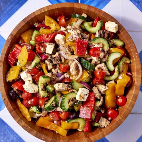 A wooden bowl filled with Greek Chicken salad—cucumbers, tomatoes, red onion, bell peppers, olives, feta cheese, and grilled chicken—rests on a blue-and-white tiled surface.
