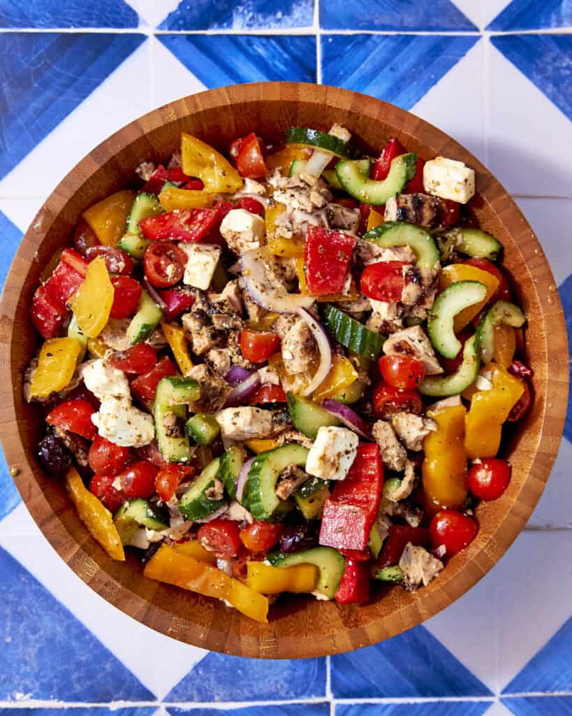 A wooden bowl filled with Greek Chicken salad—cucumbers, tomatoes, red onion, bell peppers, olives, feta cheese, and grilled chicken—rests on a blue-and-white tiled surface.