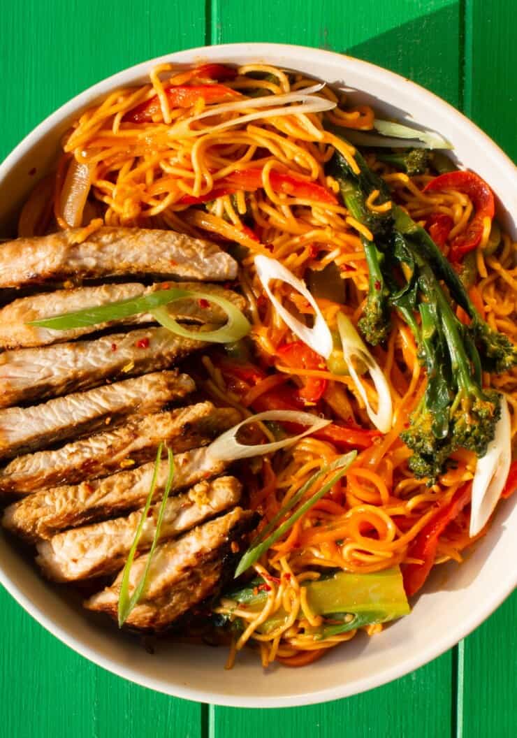A white bowl filled with noodles, sliced turkey steaks, spiralised carrots, red peppers and broccoli on a green background.