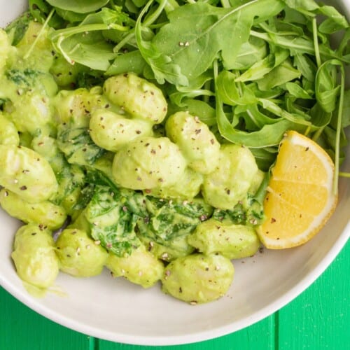 Spinach Pesto Gnocchi with Rocket with a wedge of lemon in a white bowl on a green background.
