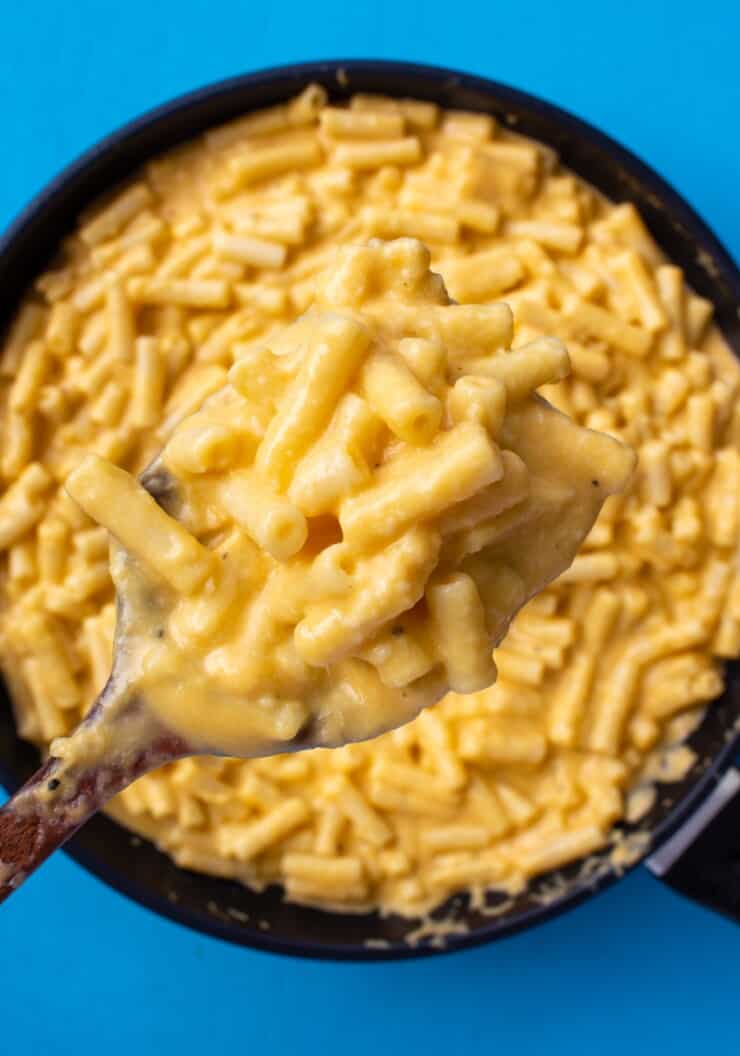 A large pan of creamy Mac and cheese with a serving lifted up on a wooden spoon with a blue background.