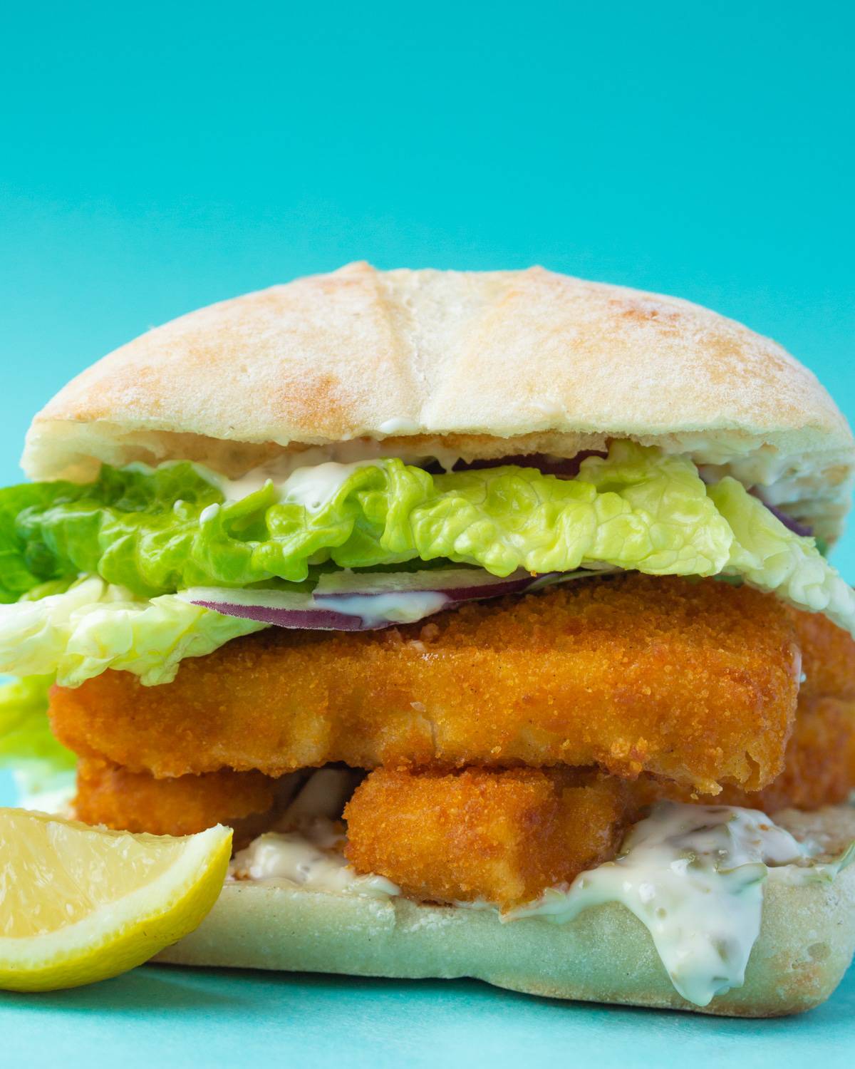 Side view of fish finger sandwich with lettuce, red onion, mayonnaise and a wedge of lemon with blue background.