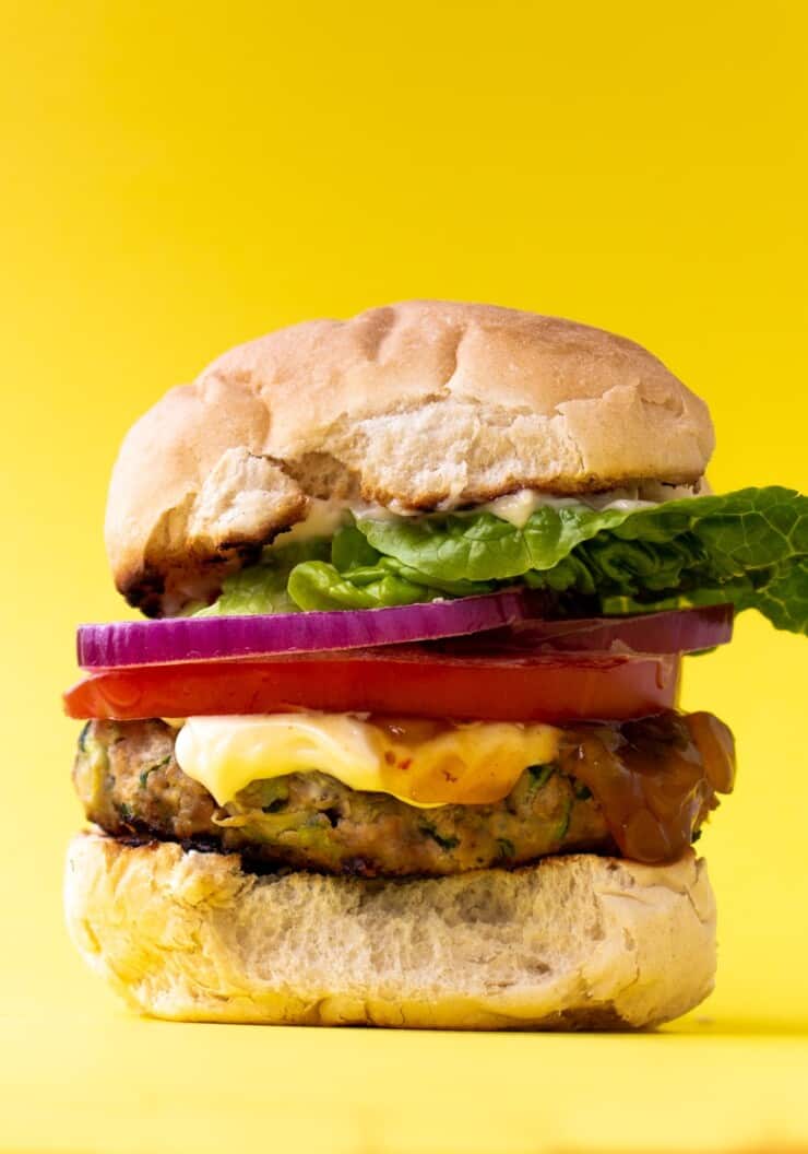 Side view of turkey burger with lettuce, red onion, mayonnaise and tomato in a bun with a yellow background.