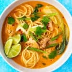 A bowl of chicken and noodle soup topped with coriander, lime wedges and chilli flakes.