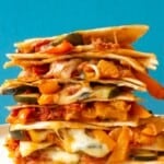Side view of vegetable quesadillas piled high with roasted vegetables, tortillas and cheesy filling with a blue background.