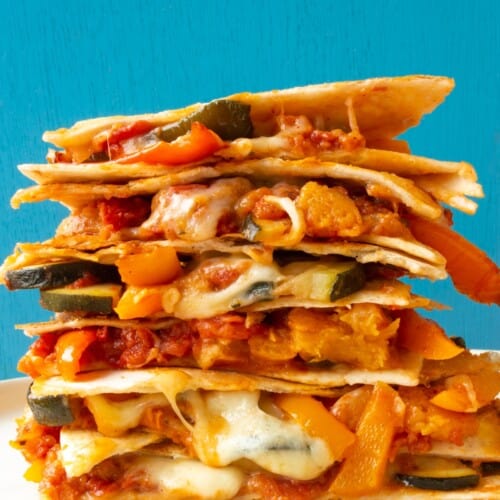 Side view of vegetable quesadillas piled high with roasted vegetables, tortillas and cheesy filling with a blue background.