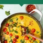 Top-down view of cod curry in coconut milk, garnished with cherry tomatoes, greens, and red chili flakes. A wooden spoon rests in the flavorful cod curry, with a bowl of chili flakes beside the pot for added heat.