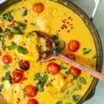A skillet filled with cod curry in yellow coconut milk sauce, cherry tomatoes, spinach, and red chili flakes. A wooden spoon lifts a portion. Text reads “Cod Curry with Coconut Milk” and “Quick One-Pan Dinner.” Perfect for easy cod curry nights.