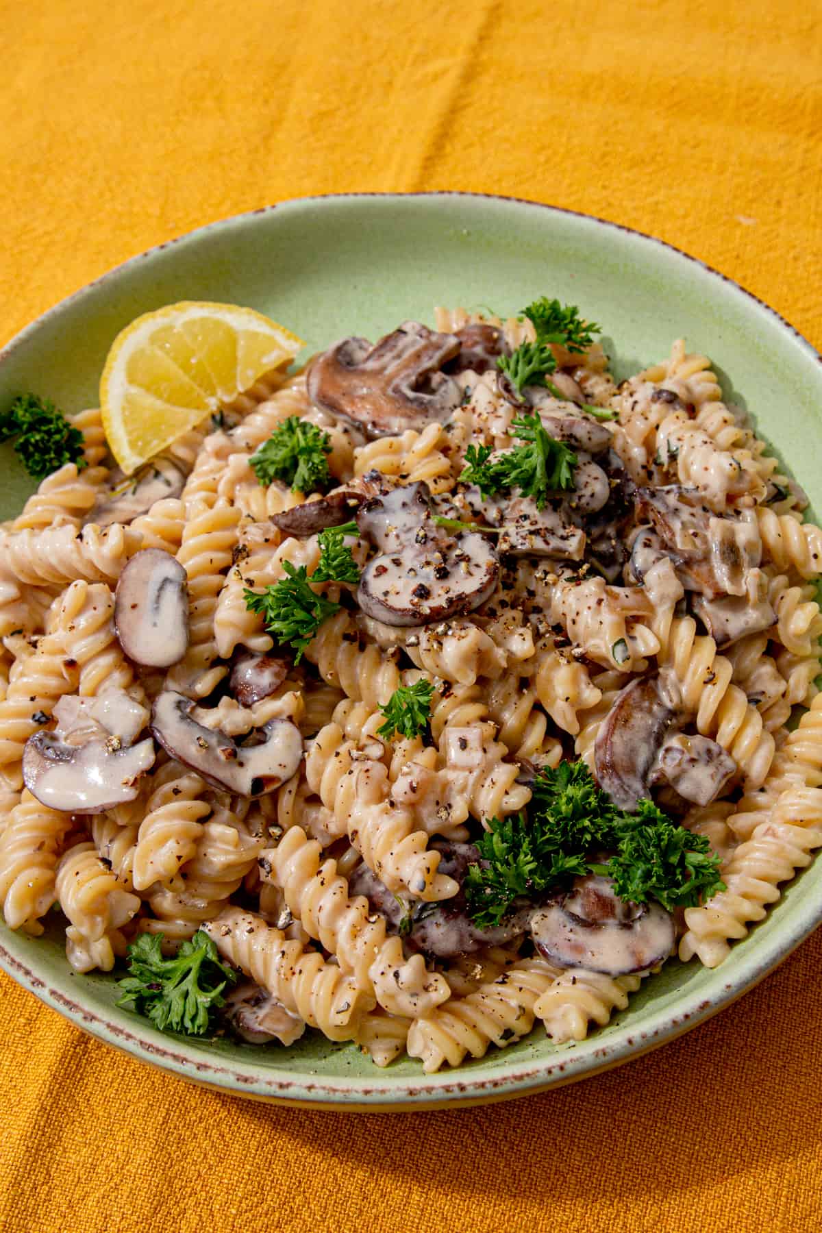 A pale green bowl with pasta and mushrooms in a sauce with a wedge of lemon.