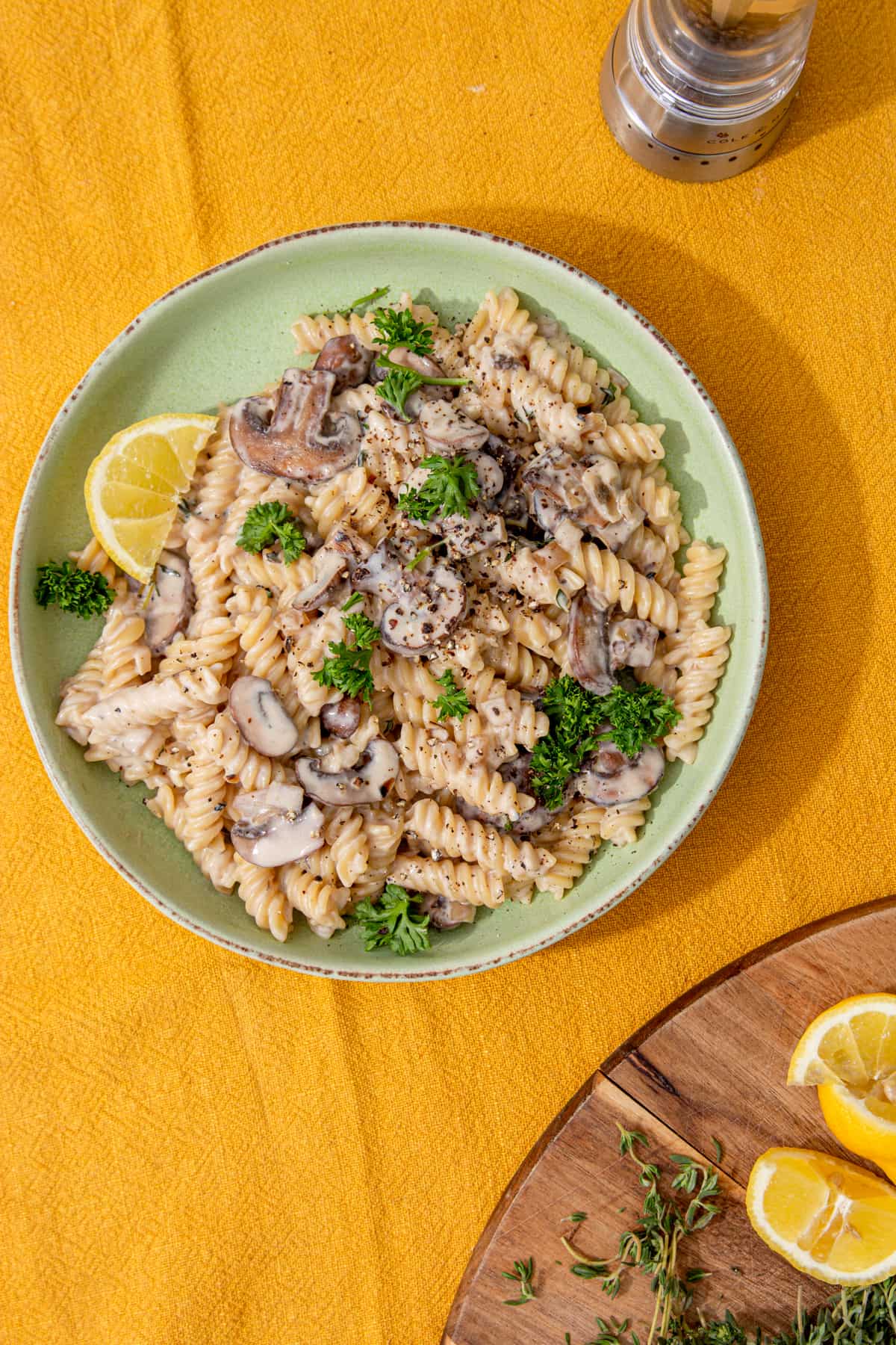 A bowl of pasta and mushrooms in a sauce with a wedge of lemon and a copping board with lemon wedges.