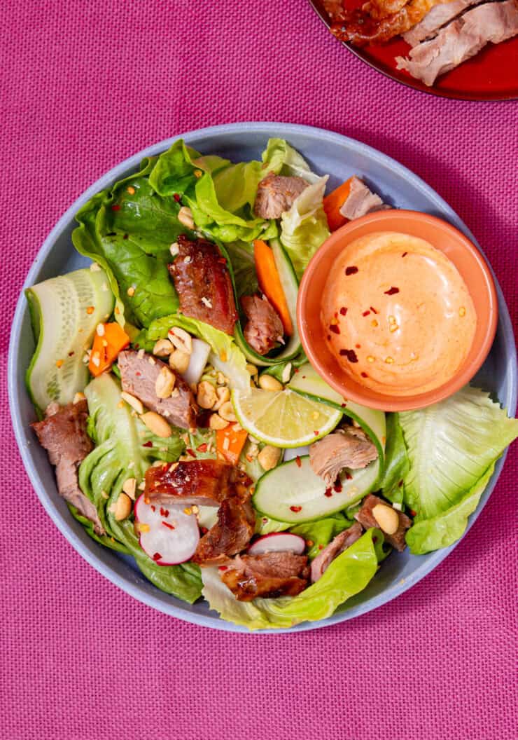 A bowl of salad with turkey pieces, a lemon wedge and a small bowl of sriracha/mayo sauce on a pink background .