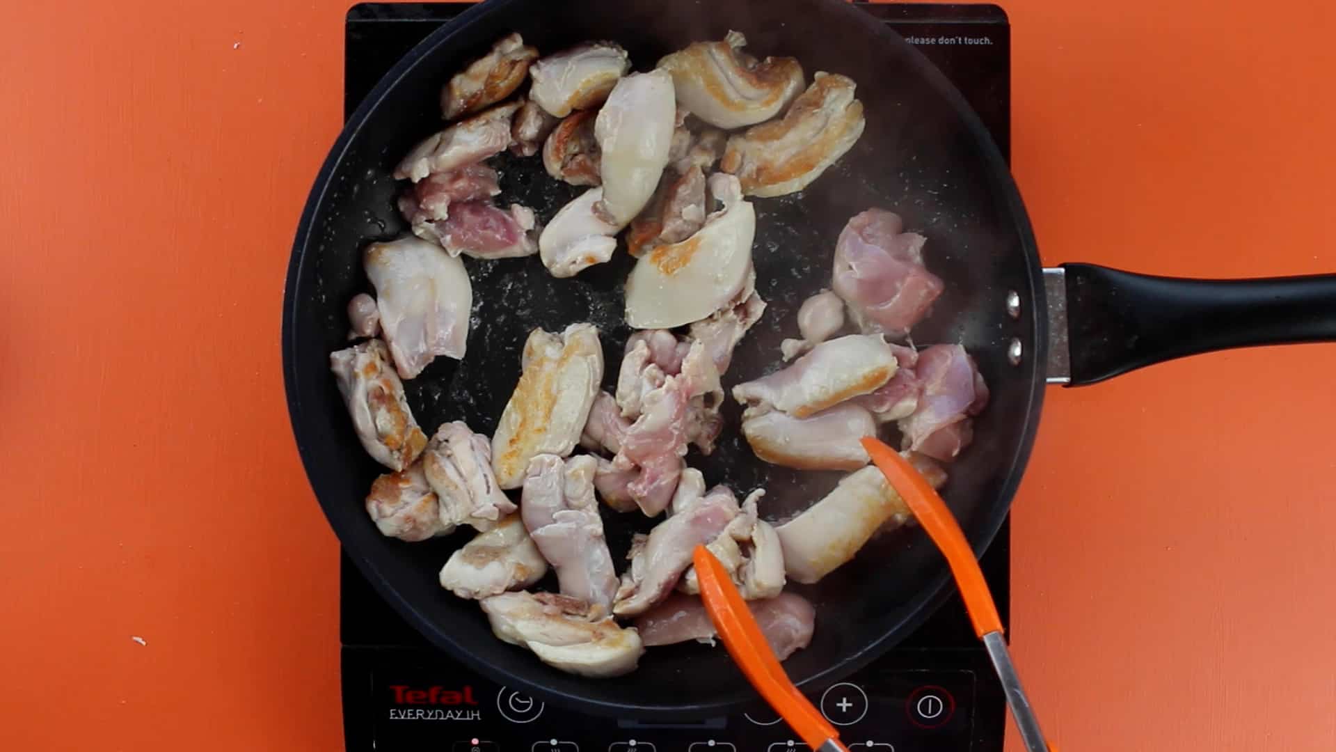 Chicken pieces frying in a pan and rotated with orange tongs on a stove on an orange background.